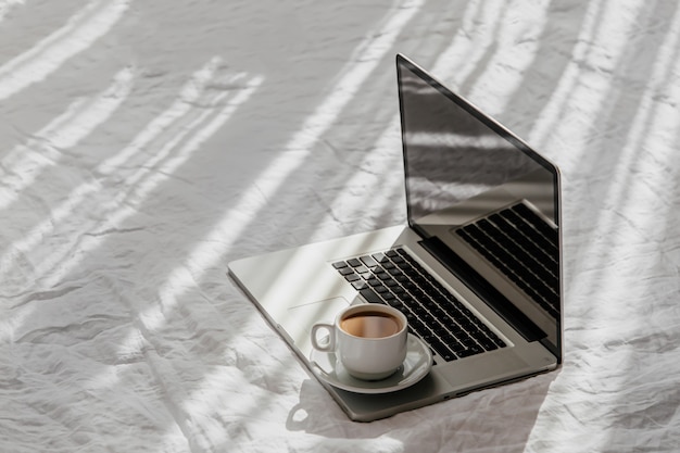 Resta a casa, in quarantena. Lavorare da casa. Computer portatile e tazza di caffè sul letto bianco. Concetto di lavoro a casa. Luce del mattino . concetto di stile di vita