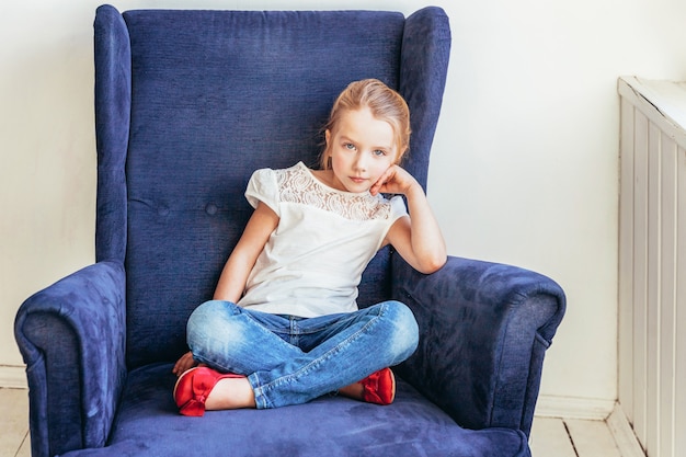 Resta a casa Resta al sicuro. Bambina dolce in jeans e maglietta bianca che si siedono sulla sedia blu accogliente moderna che si rilassa nel salone luminoso bianco a casa all'interno. La gioventù degli scolari dell'infanzia rilassa il concetto.