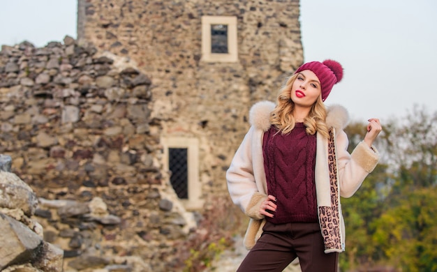 Resta caldo e alla moda. La donna indossa un cappotto di pelliccia. Abiti invernali. Armadio per la stagione fredda. La ragazza sta vicino alle rovine di pietra. Cappotto fantasia chic. Cappotto in montone di lana naturale. Pelliccia sul cappuccio. Colpo di stagione.