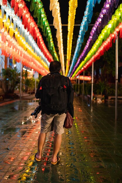 Retro dell'uomo asiatico zaino in spalla che tiene la macchina fotografica che cammina in un pedone decorato con lanterna di carta illuminata colorata nella tradizione annuale di appendere la lanterna nel nord della Thailandia di notte a Lamphun