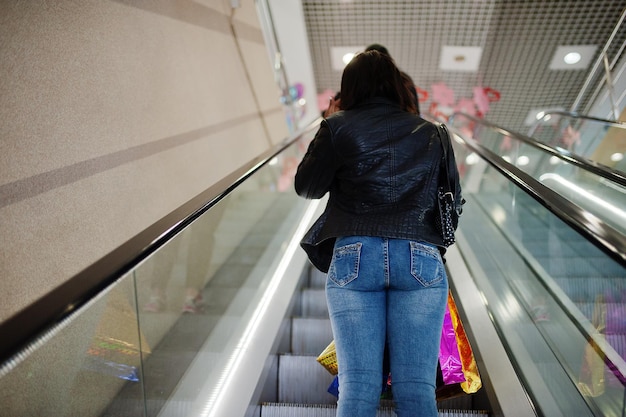 Retro della donna afroamericana che cammina con le borse della spesa colorate nel centro commerciale del centro commerciale sulla scala mobile.