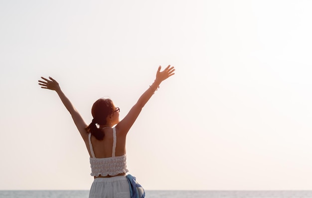 Retro della donna asiatica con occhiali da sole felice alza le mani in alto posa sulla spiaggia in vibrazioni estive Felice donna in canotta bianca che guarda il cielo sul mare e cielo aperto con spazio copia Vacanze