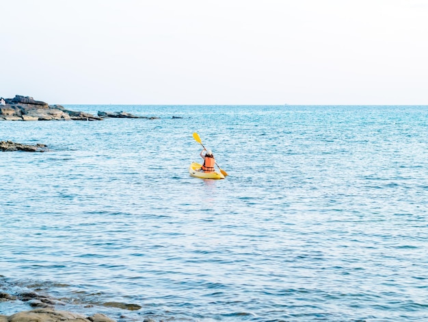 Retro della donna asiatica in giubbotto di salvataggio e berretto in kayak su kayak giallo con l'utilizzo della pagaia sul mare Femmina felice che si diverte attività su sfondo vista mare Vacanza viaggio vacanza