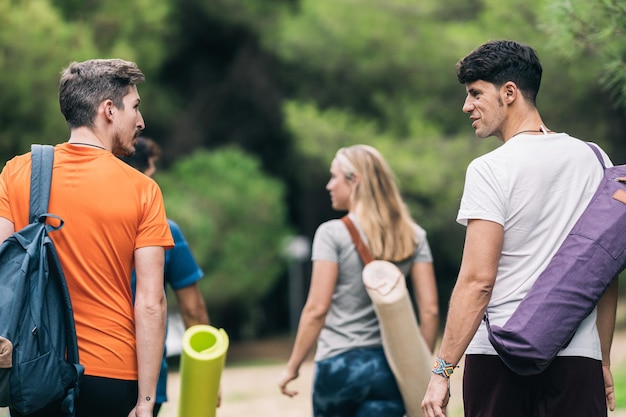 Retro di persone che chiacchierano mentre tengono in mano stuoie e borse per lo yoga e passeggiano nella foresta