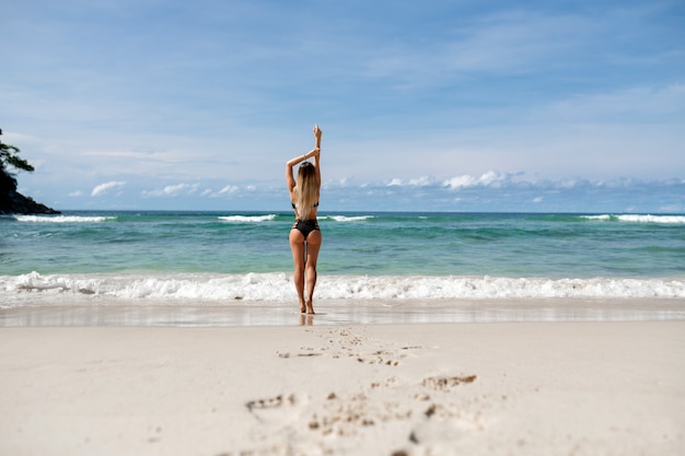 Retrovisione: bionda, bella ragazza che posa sulla spiaggia e che alza le braccia. Vacanza tropicale. Viaggia in paesi caldi