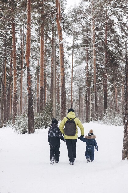 Retrovisione del padre con lo zaino e i figli piccoli che si tengono per mano camminando insieme nella foresta innevata d'inverno. Attività invernale all'aperto. Concetto di viaggio locale e fine settimana in famiglia