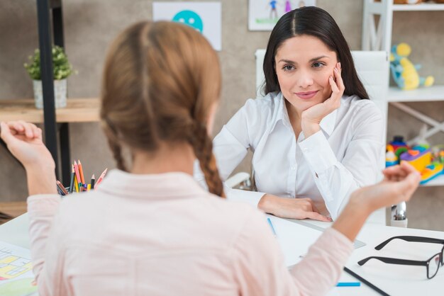 Retrovisione della bambina che parla con suo psicologo femminile sorridente nell&#39;ufficio