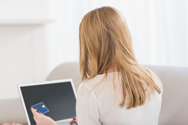 Retrovisione di una bionda graziosa che compera online con il computer portatile