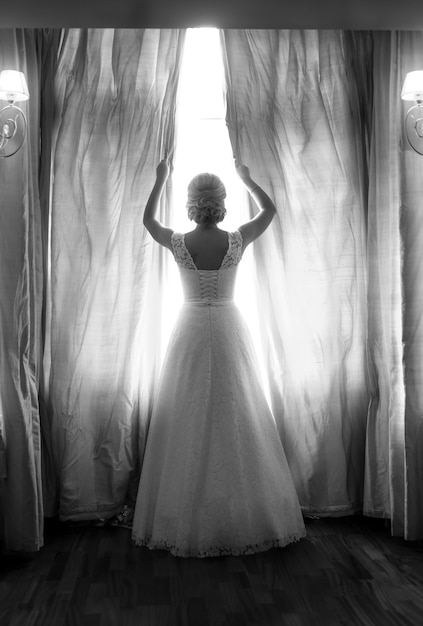 Retrovisione in bianco e nero della sposa elegante che posa alla grande finestra in camera da letto