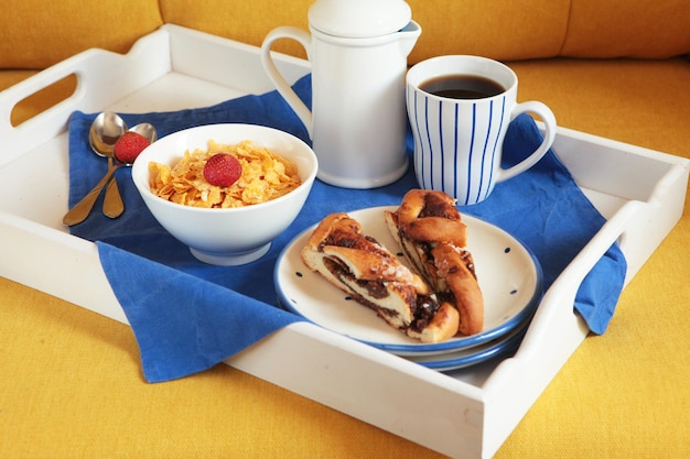 Ricca colazione continentale su una tavoletta di legno bianca e un divano giallo. Cornetti croccanti francesi, muesli, frutti di bosco dolci e caffè caldo per gustosi pasti mattutini. Delizioso inizio di giornata.