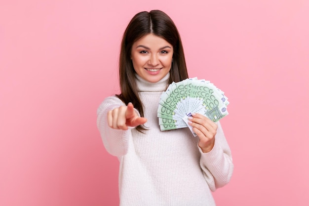 Ricca giovane donna felice che tiene banconote in euro e punta il dito verso la fotocamera, scegliendo te come vincitore, indossando un maglione bianco in stile casual. Studio indoor girato isolato su sfondo rosa.