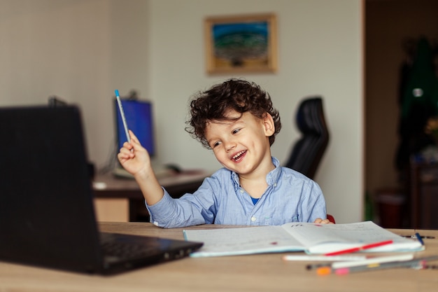 Ricci ragazzo carino a casa formazione online un bambino in età prescolare a un computer portatile impara e scrive in un taccuino un compito