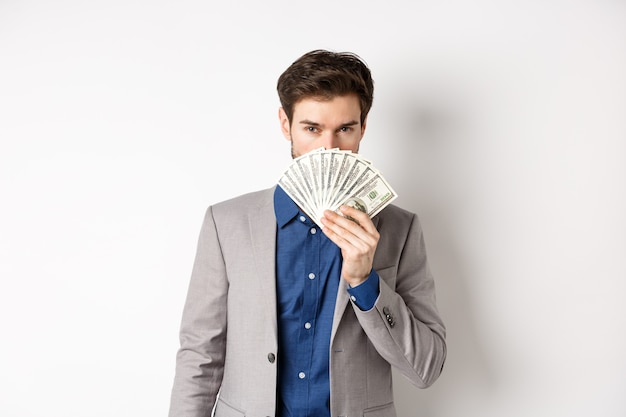 Ricco uomo d'affari in tuta che mostra banconote da un dollaro, nascondendo la faccia dietro i soldi con lo sguardo sensuale alla macchina fotografica, sfondo bianco.