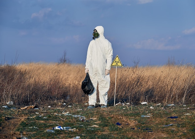 Ricercatore in tuta protettiva che raccoglie immondizia di plastica nel sacco nero dei rifiuti all'aperto in una giornata di sole