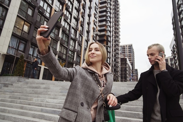 Ricorda questo momento! Giovane donna che tiene smartphone e prendendo selfie con il suo fidanzato. Giovani coppie che camminano nella città di autunno. Blocchi di appartamenti sullo sfondo. Uomo che parla al telefono.