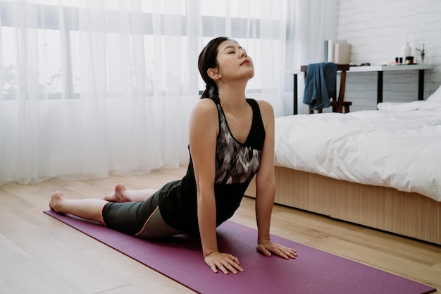 ricreazione al coperto donna asiatica che fa yoga per liberare lo stress in camera da letto. ragazza cinese che pratica la posa del cobra raddrizzando le braccia per sollevare il petto dal pavimento, chiudendo gli occhi e inspirando profondamente