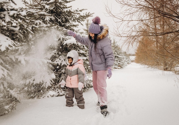 Ricreazione invernale all'aperto. La madre felice si scrolla di dosso la neve da un ramo su un bambino in piedi tra gli abeti innevati