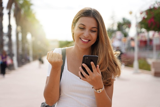 Ridendo eccitato giovane donna guardando il suo smartphone celebrando all'aperto alzando il pugno in esultanza