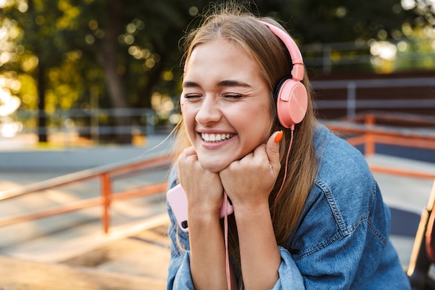 ridendo felice positivo giovane adolescente fuori nel parco ascoltando musica con le cuffie che tengono il telefono cellulare che mostra la lingua.