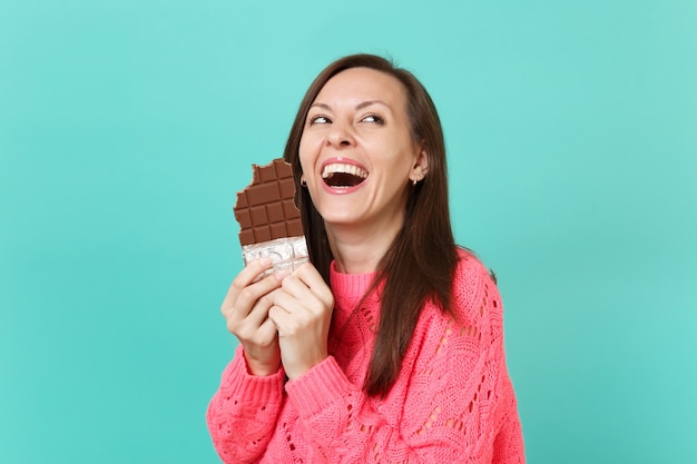 Ridendo giovane donna in maglia maglione rosa guardando in alto, tenendo in mano mangiare barretta di cioccolato isolato su sfondo blu muro turchese, ritratto in studio. Concetto di stile di vita della gente. Mock up copia spazio.