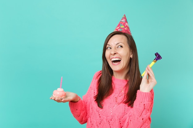 Ridendo giovane donna in maglia rosa maglione, cappello di compleanno con la riproduzione di tubo cercando tenendo in mano la torta con la candela isolata sul fondo della parete blu. Concetto di stile di vita della gente. Mock up copia spazio.