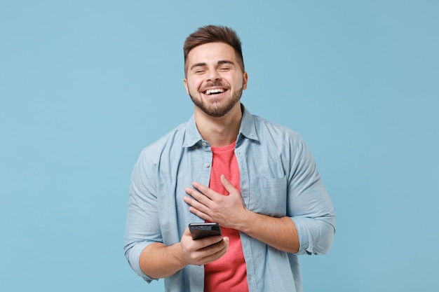 Ridendo giovane ragazzo barbuto in camicia casual in posa isolato su sfondo blu pastello parete ritratto in studio. Concetto di stile di vita delle emozioni delle persone. Mock up spazio di copia. Usando il telefono cellulare, metti la mano sul petto.