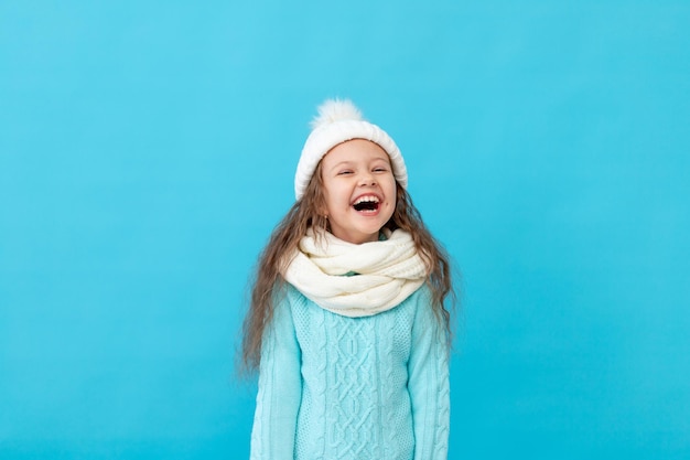 Ridere bambina in abiti invernali cappello e maglione su uno sfondo blu isolato sorridente e divertirsi, un luogo o uno spazio per il testo