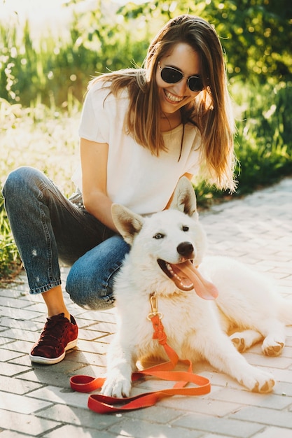 Ridere bella giovane donna in abiti estivi e occhiali da sole seduto su hunkers con il suo cane bianco felice