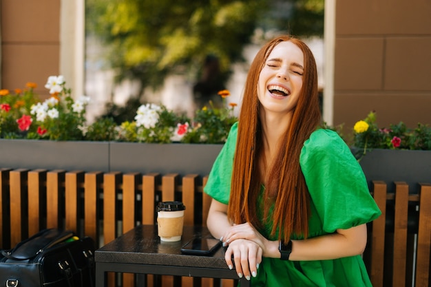 Ridere giovane donna seduta a tavola con una tazza di caffè e telefono cellulare nella terrazza del caffè all'aperto