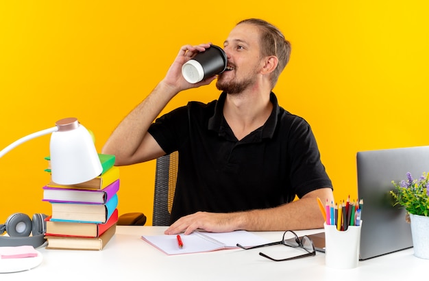 Ridere giovane studente seduto a tavola con gli strumenti della scuola beve caffè