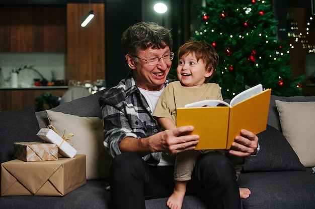 Ridere nonno e suo nipote seduti su un divano guardando attraverso un album di foto. Il bambino guarda un vecchio. Albero di Natale sullo sfondo.