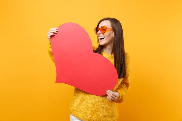 Ridere piuttosto giovane donna in maglione di pelliccia e occhiali da vista cuore arancione che tengono cuore rosso vuoto vuoto isolato su sfondo giallo brillante. Persone sincere emozioni, concetto di stile di vita. Zona pubblicità.