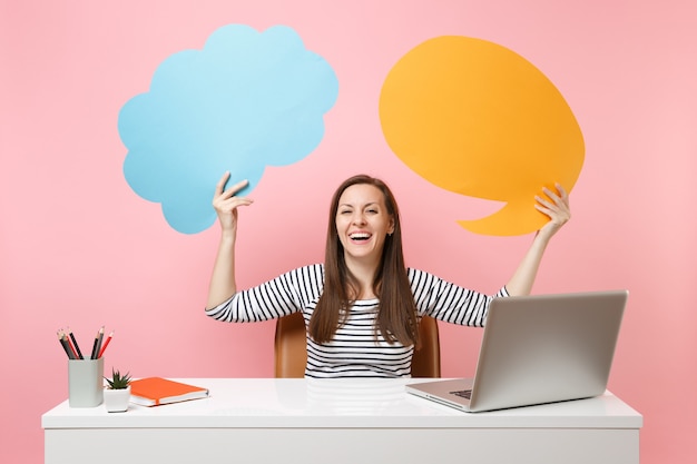 Ridere ragazza tenere blu giallo vuoto vuoto Say cloud nuvoletta lavoro alla scrivania bianca con laptop isolato su sfondo rosa pastello. Concetto di carriera aziendale di successo. Copia spazio per la pubblicità.