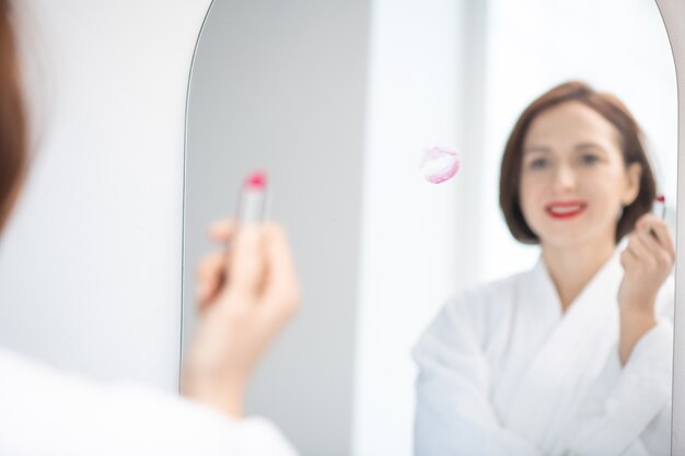 Riflessione di giovane donna in accappatoio bianco che tiene il rossetto cremisi mentre levandosi in piedi davanti allo specchio con la stampa delle labbra