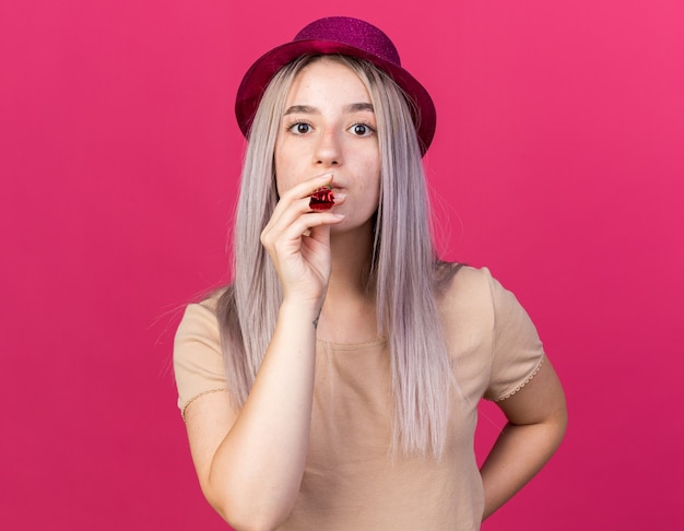 Rigorosa giovane bella ragazza che indossa cappello da festa che soffia fischio di festa mettendo la mano sull'anca isolata sulla parete rosa