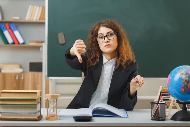 rigoroso che mostra i pollici verso il basso giovane insegnante femminile che indossa occhiali punta alla fotocamera con il puntatore seduto alla scrivania con strumenti scolastici in classe