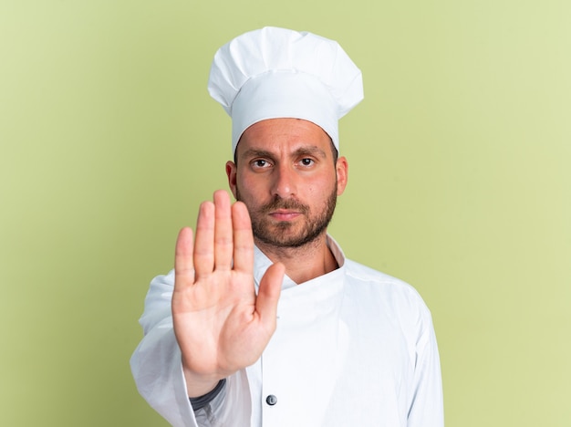 Rigoroso giovane maschio caucasico cuoco in uniforme da chef e berretto che guarda la telecamera facendo un gesto di arresto isolato sulla parete verde oliva