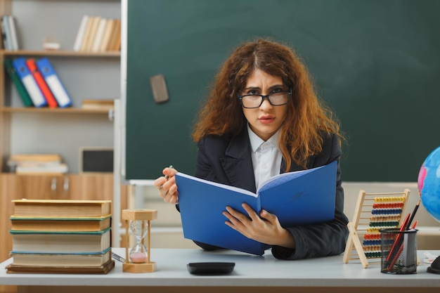 rigoroso guardando la fotocamera giovane insegnante femminile che indossa occhiali tenendo il libro seduto alla scrivania con strumenti scolastici in classe