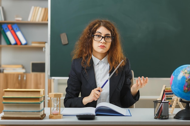 rigoroso guardando la fotocamera giovane insegnante femminile che indossa occhiali tenendo il puntatore seduto alla scrivania con strumenti scolastici in classe