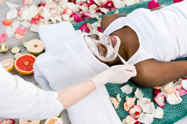 Rilassamento di riposo della bella ragazza africana nel ricorso di stazione termale con gli occhi chiusi mentre il cosmetologo applica la maschera facciale sul suo fronte.