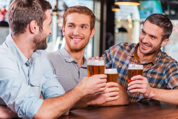 Rilassante dopo il lavoro. Tre giovani uomini felici in abbigliamento casual che parlano e bevono birra seduti insieme al bancone del bar