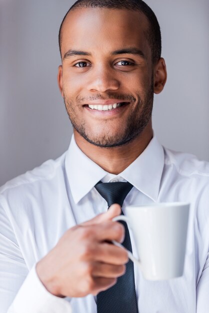 Rilassante durante la pausa caffè. Bel giovane africano in camicia e cravatta che tiene tazza con bevanda calda