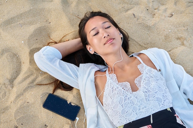 Rilassante giovane donna in cuffia con smartphone sulla sabbia vista dall'alto