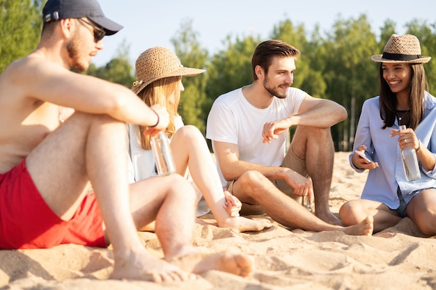 Rilassarsi con i migliori amici. Gruppo di giovani felici seduti insieme in spiaggia a parlare e bere birra