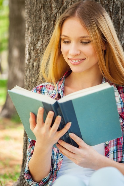 Rilassarsi con un buon libro. Bella giovane donna che legge un libro e sorride mentre si appoggia all'albero in un parco