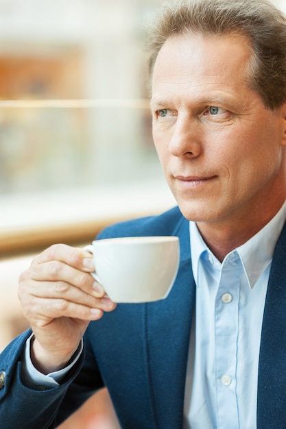 Rilassarsi con una tazza di caffè fresco. Uomo maturo premuroso in abiti da cerimonia che beve caffè e guarda lontano mentre è seduto in una caffetteria