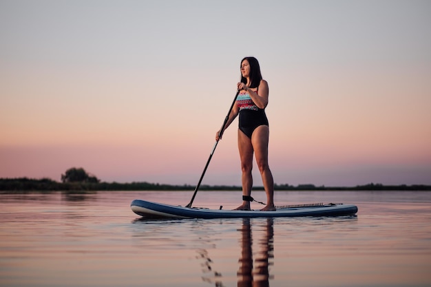 Rilassata donna di mezza età che rema a bordo sup con remo guardando il tramonto sul lago acqua increspata con cielo rosa sullo sfondo Stile di vita attivo per le persone anziane