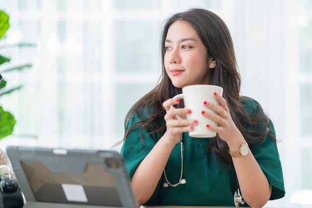 Rilassatevi dopo il lavoro asiatico femminile medico tenere in mano la tazza della bevanda del caffè caldo casual rilassarsi dopo aver lavorato duramente in telemedicina in clinica mentre indossano l'uniforme