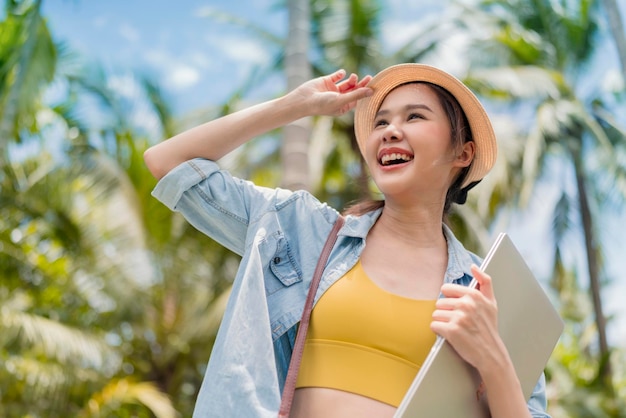 Rilassati casual spensierato sorridente asiatico femmina nomade digitale programmatore freelance tenere in mano laptop camminare sulla spiaggia e albero di cocco blu skynew stile di vita millenario lavorando ovunque con felicità