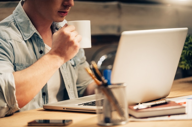 Rimanere concentrato. Primo piano di un giovane fiducioso che lavora fino a tardi e beve caffè mentre è seduto al suo posto di lavoro